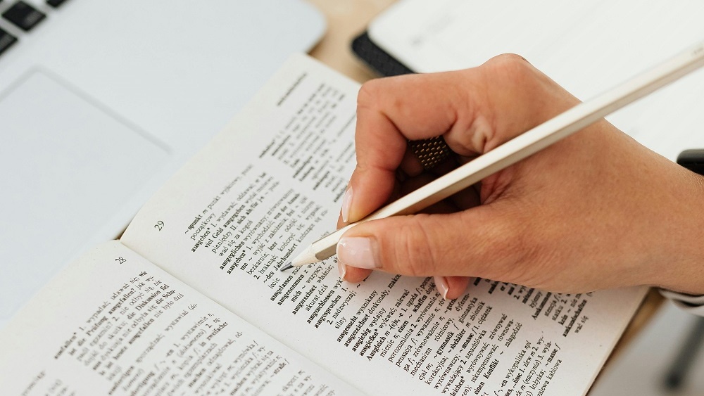 Close-up of a person’s hand using a pencil to underline text in a dictionary, with a laptop in the background, used in a vocabulary quiz created with Opinion Stage.